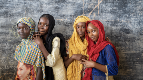 Four girls standing in front of a chalkboard, looking at the camera with their arms around one another. JRS programs for women and girls seek to empower them.